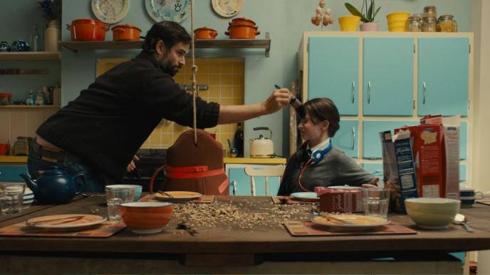 A man paints the front of a teenage girl's head with a paint brush, in the kitchen