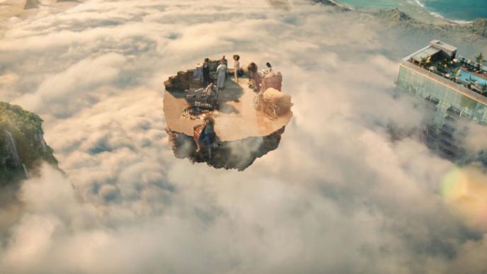 A floating island with people on it surrounded by clouds. On the left there is the roof of a holiday resort.