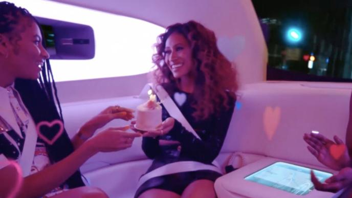 Three women sitting in a futuristic car one is holding a birthday cake.