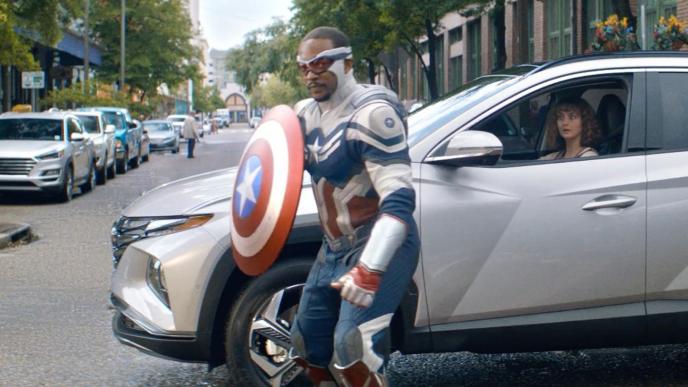 Captain America holding his shield standing in front of a silver car in the middle of the street.