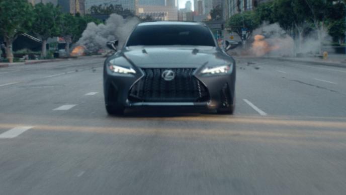 A car is driving towards the camera down a city street with explosions in the background.