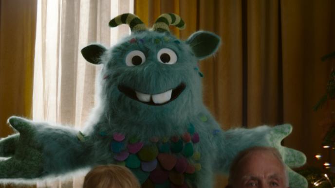 A blue fuzzy monster with stripey horns is standing in a living room, there are the foreheads of three people in the foreground.