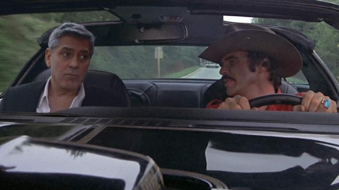 Two men in a car the man driving is wearing a hat and looking at the man in the passenger side.