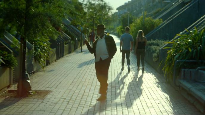 A man dancing down a pedestrian street with trees and plants to the side. There are people walking on the street behind him.