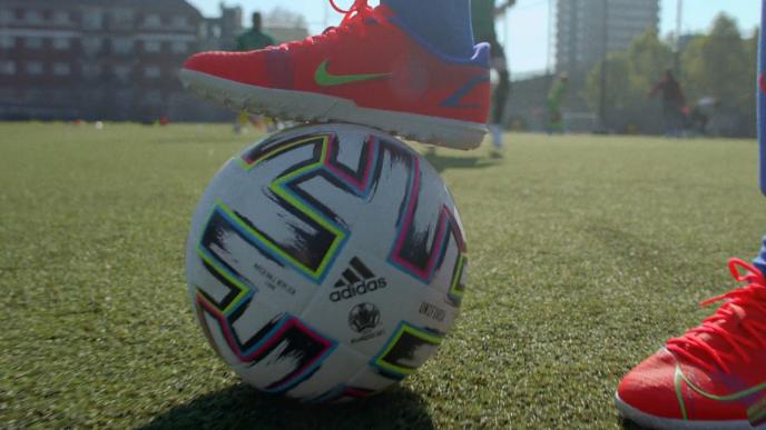 A pair of feet in orange trainers, one of the feet is on top of a football on a football pitch.