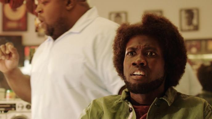 A man with a panicked look on his face faces out towards the camera, there is a man with a white shirt in the background.