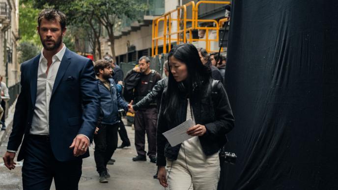 A man and a woman are walking down a street on a film set with many other people in the background.