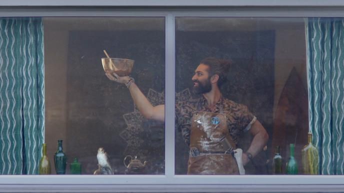 A man is seen through a window. He wears a work apron and proudly holds a bowl of batter