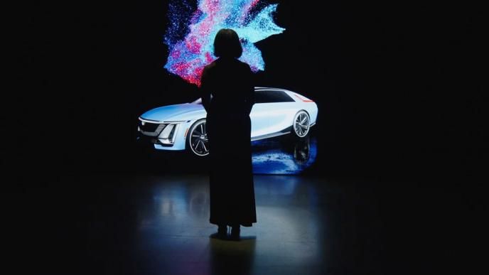 The silhouette of a person as they stand in front of a large screen in a dark room. On the screen is the Cadillac Lyriq car