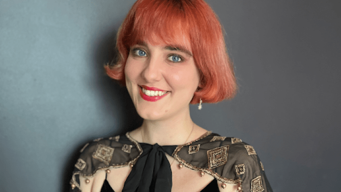 A woman wearing a black dress with red hair and red lipstick against a black background.