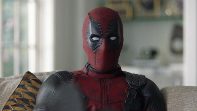 Deadpool is sat on a sofa looking at the camera with his feet up.