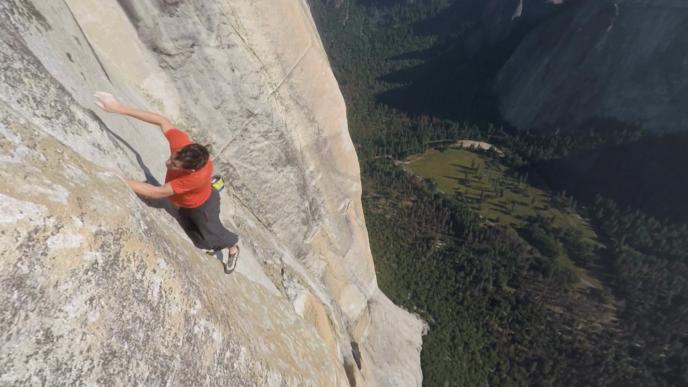 A man free climbing a large rock face above a forested hillside.