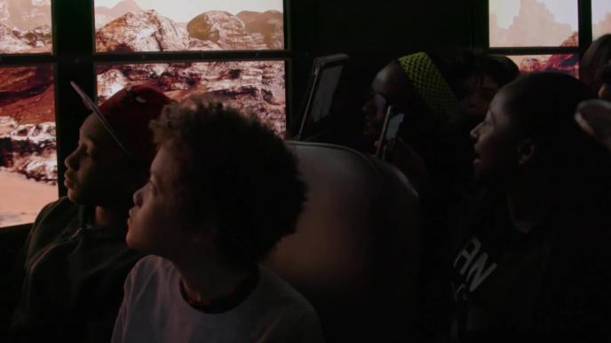 A group of school children in a school bus look out the bus windows into what looks like the planet Mars. The windows are replaced by screens which create the effect that the bus is driving on Mars