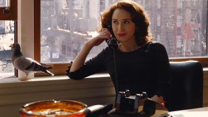 Miriam 'Midge' Maisel sits at a desk chair with a cityscape behind her seen through the windows