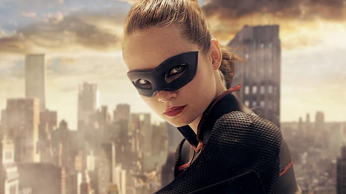 A woman with a superhero mask  standing on the top of a building with a cityscape in the background.