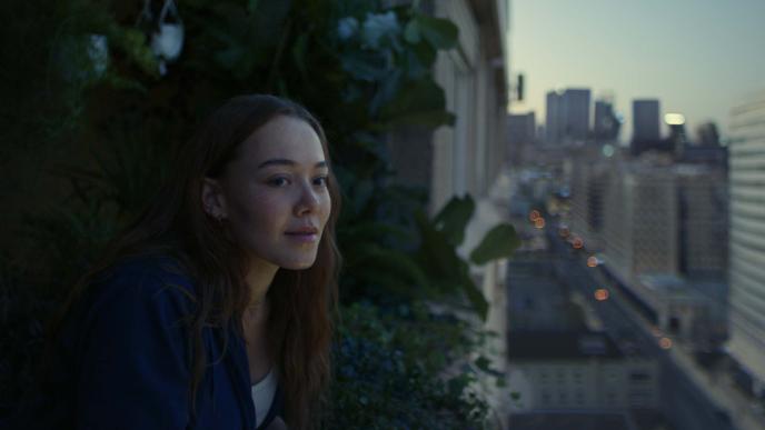 Concrete block of flats focusing on one balcony full of green plants whilst a woman looks out into the distance