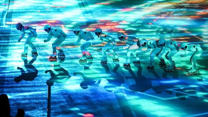 Eight people wearing white snowsuits are ice skating in a line from right to left. There are blue and green lights projected on the ice. 