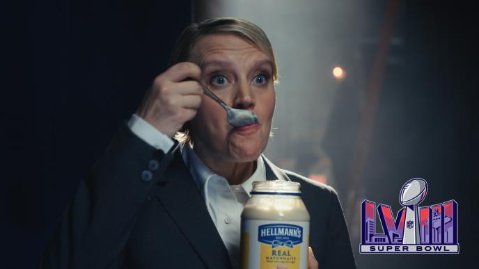 A woman on a stage in a suit eating mayonnaise from a jar, there is the Super Bowl LVIII logo in the bottom left corner.