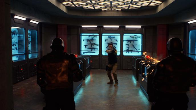 A man in body armour pointing a gun at two soldiers with their backs to the camera, there are many more weapons in cabinets behind the man. 