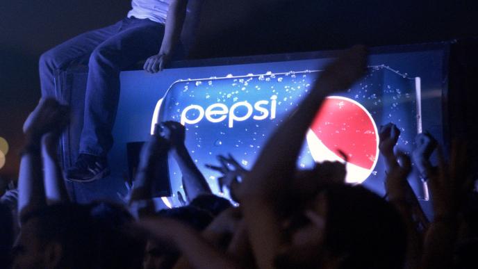 A Pepsi vending machine crowd surfs through a crowd, someone sits on top of the vending machine