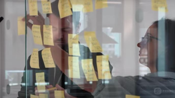 Two people writing on yellow sticky notes and sticking them to a glass wall