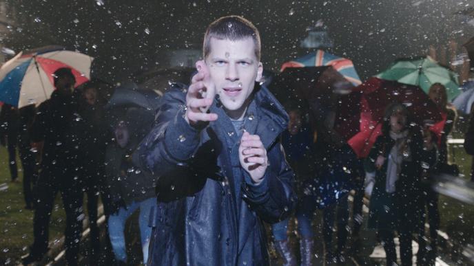 actor jesse eisenberg as j daniel atlas holding his hands in front of his chest as he is conjuring water droplets in the air. there are people with umbrellas walking by