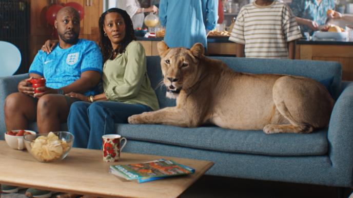 A lioness lays on a living room couch as a couple looks shocked sitting next to it