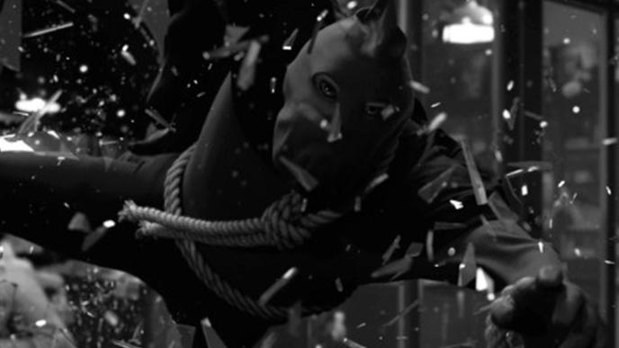 A man wearing a black outfit and facemask is jumping through a window.