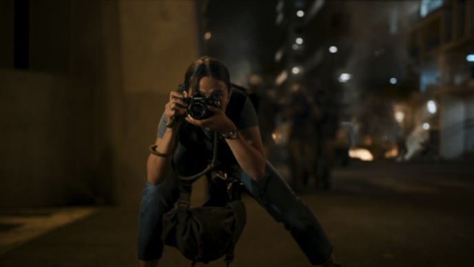 A woman holding a camera and wearing a bulletproof vest is crouching and taking a picture in front of a US street in the dark.