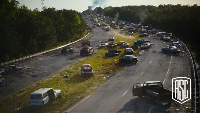 A destroyed highway with many broken down cars and fire in the background.