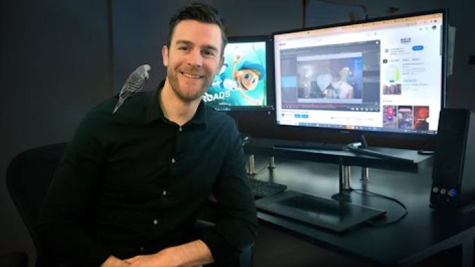A photograph of a man sat in front of a computer with a blue budgie on his shoulder.