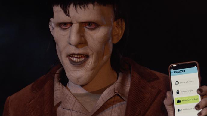 A Frankenstein-esque monster holds up a cell phone with the GEICO app displayed