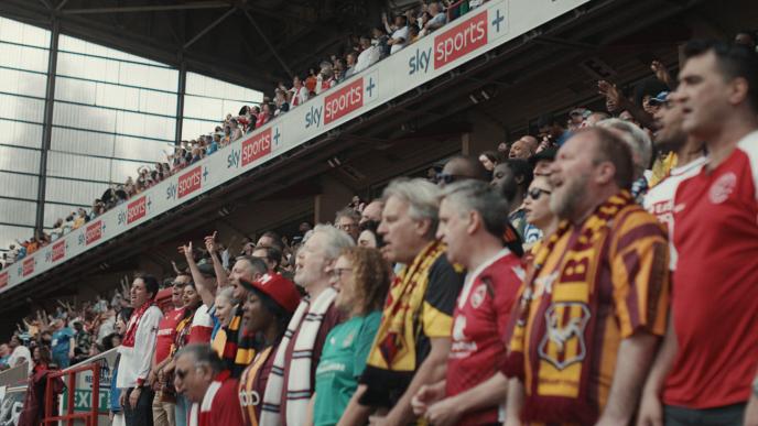 A crowd of football fans standing in the stands of the stadium with the Sky Sports banner above their heads. 