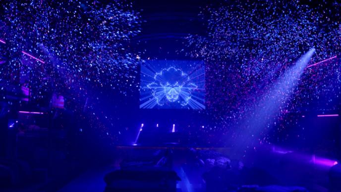 a dark dancefloor that is lit with spotlights from the right top corner. there is confetti and glitter in the air. there is a screen in the top centre that has an outline of a sheep 