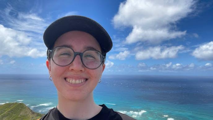 A woman wearing glasses and a hat smiling at the beach with a blue sky and clouds behind her.