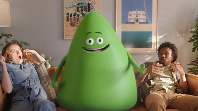 A large green Cricket mascot sits on a couch while two people look in awe