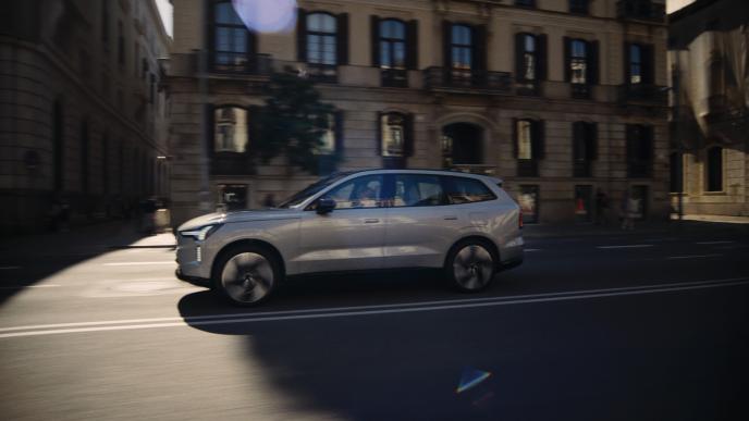 A dark silver Volvo car driving into the sunlight along a city street. 
