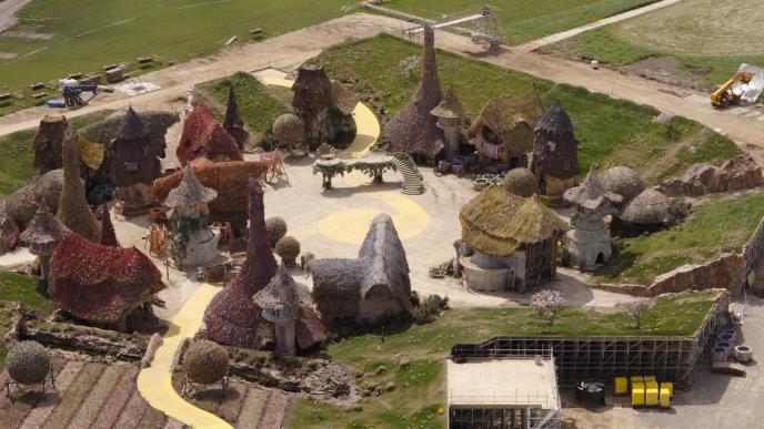 Plate footage from the set of Munchkinland in Wicked Part 1. From a distance, an empty set of cottages surrounded by manufactured hills.