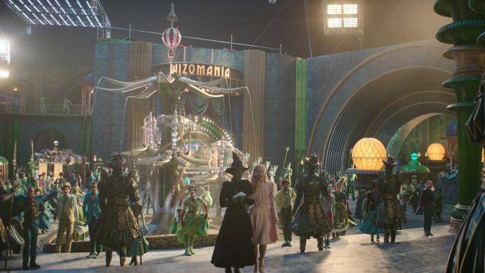 A still from the set of Wicked part one, during One Short Day. Elphaba and Glinda stand on steps, in front of a square with an elaborate fountain. Studio lights and blue screen can be seen behind the sets.