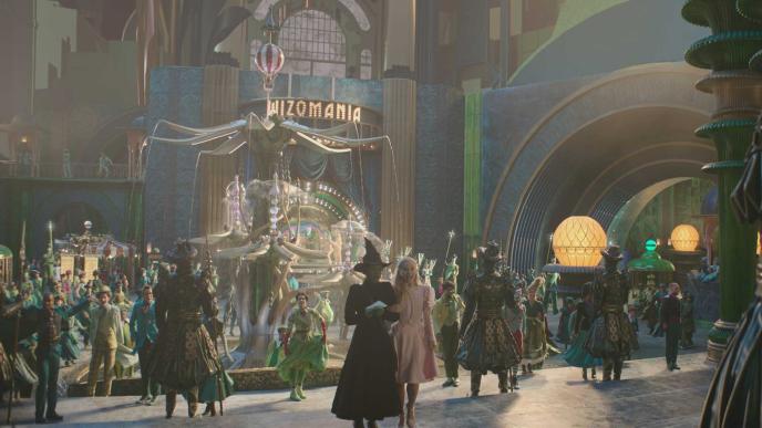 A still from the set of Wicked part one, during One Short Day. Elphaba and Glinda stand on steps, in front of a square with an elaborate fountain. Postvis has been completed to add an initial layer of animation to the sets, extending them to the full height of the shot.