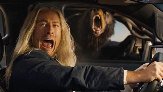 Glen Powell with long blonde flowing hair drives a truck and screams as he sees a (CG animated) bear in the vehicle next to him