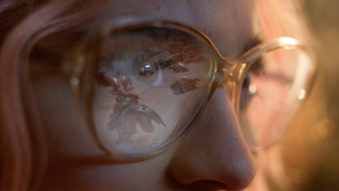 A close up on a woman's face as she plays Monster Hunter Wilds, the reflection of the game play in her glasses