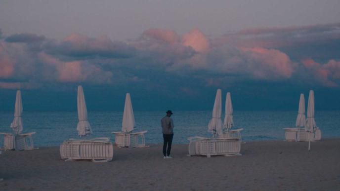 a person standing on the beach looking at the sea and mountains that are blankeed with pinky clouds. there are closed umbrellas and tables dotted around the beach