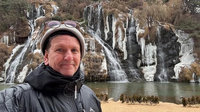 A man wearing a black coat, hat and glasses stoon in front of a frozen waterfall.