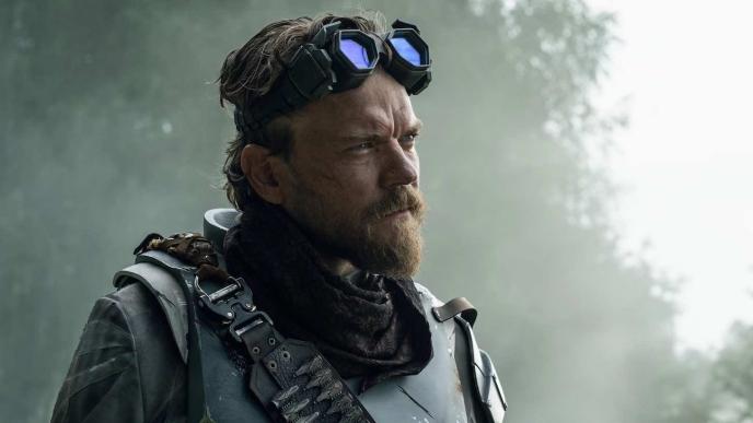 A man in post-apocalyptic fashion stands in a misty woodland