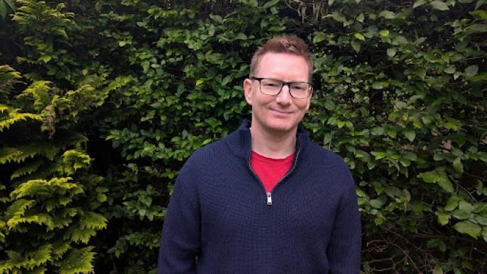 A man in a dark blue jumper, wearing glasses stood in front of a leafy background. 