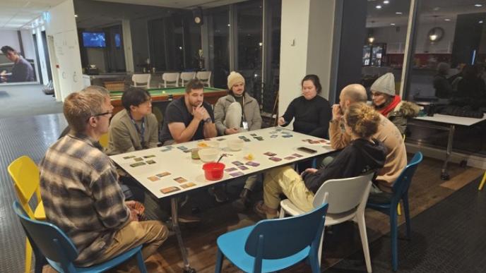 A diverse group of adults are gathered around a table playing a board game in a modern office space.