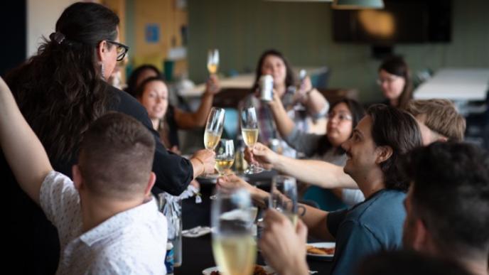 A diverse group of people are raising their glasses in a toast at a celebratory dinner with food and drinks on the table.