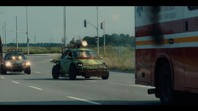 A man in a mask shoots a gun (with CG muzzle shots) while standing on a modified Slug Bug car on the interstate