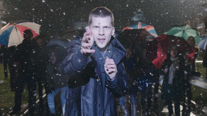 actor jesse eisenberg as j daniel atlas holding his hands in front of his chest as he is conjuring water droplets in the air. there are people with umbrellas walking by